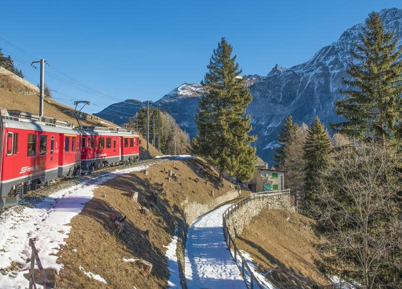 Ein roter Zug fährt eine verschneite Strecke in den Bergen hinunter.